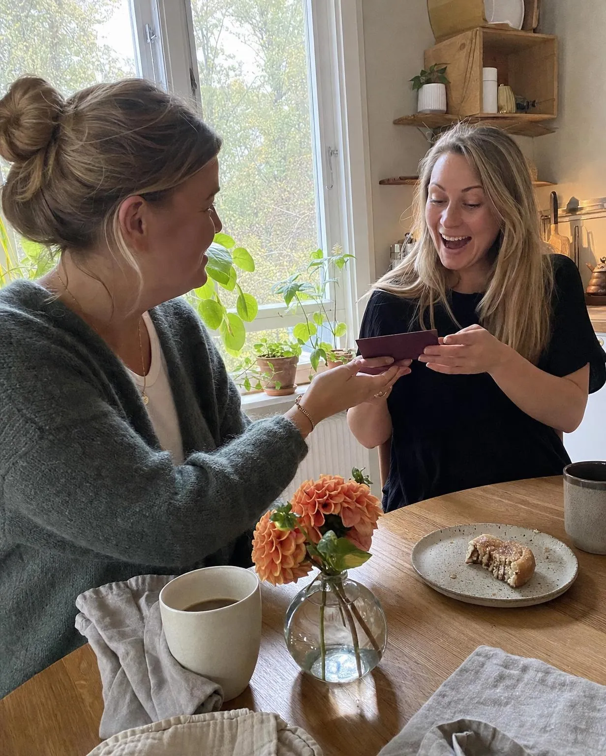 Vid ett köksbord i dagsljus räcker en kvinna sin väninna ett vinrött kuvert, med kaffe och dahlior på bordet